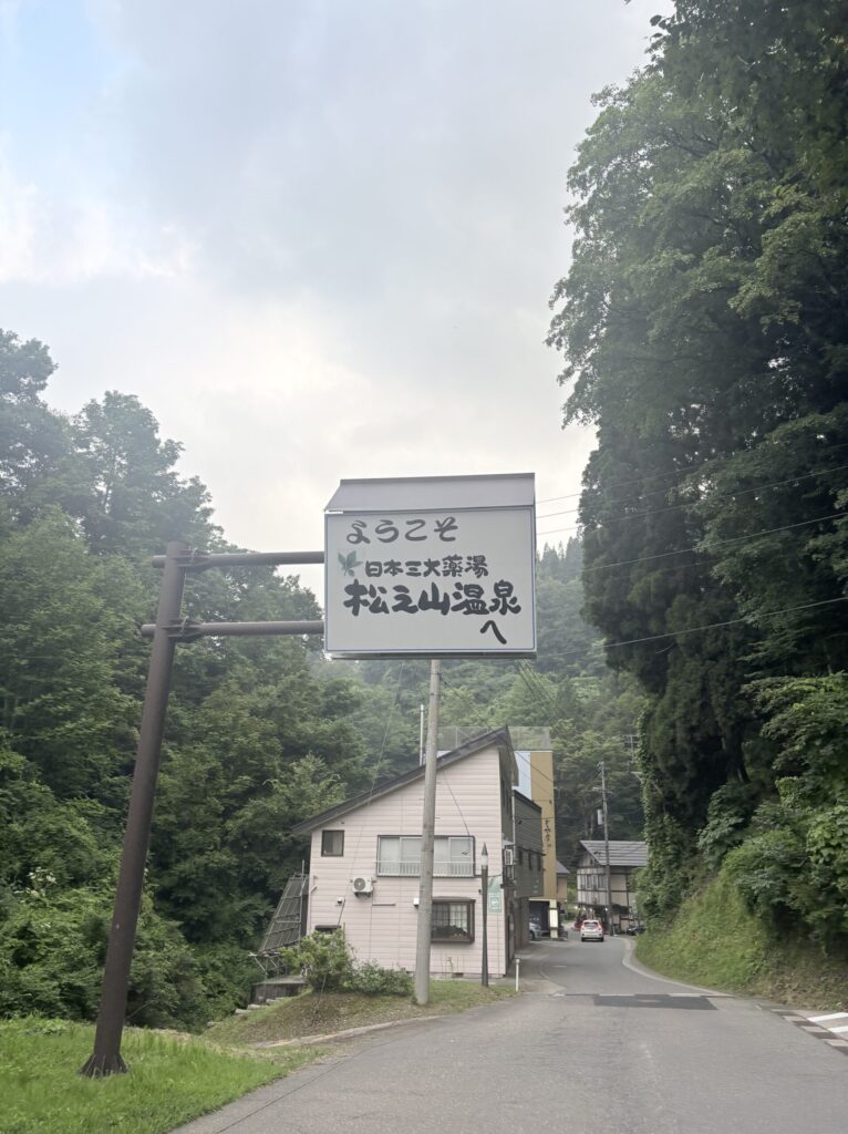 「ようこそ 日本三大薬湯 松之山温泉へ」と書かれた道路沿いの案内看板。奥に温泉街が続く