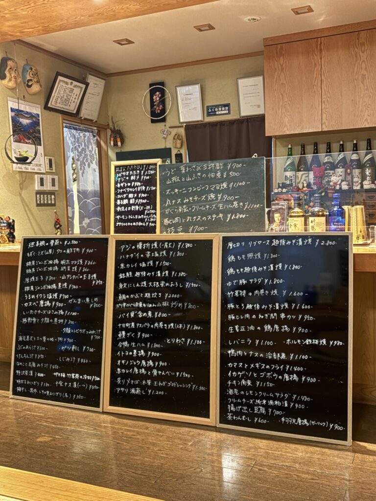 寿々木の店内。手書きの黒板メニューが3枚並び、奥の棚に日本酒の瓶が並んでいる