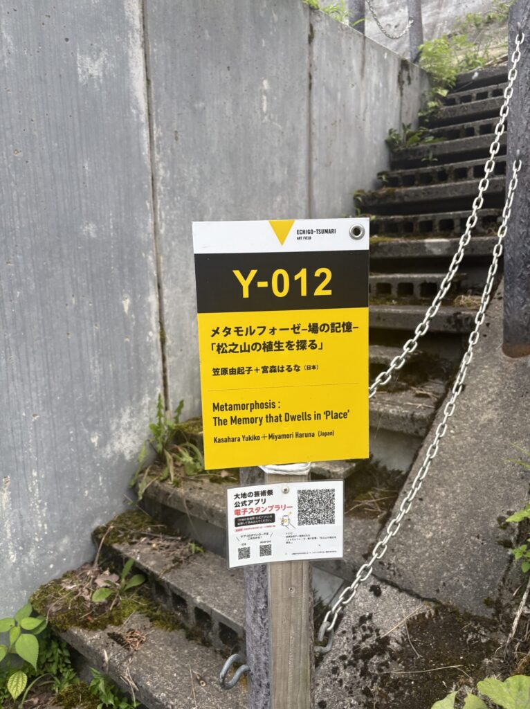 「Y-012 メタモルフォーゼ－場の記憶－『松之山の植生を探る』」と書かれた黄色い作品標識。奥にブロックの階段が続く